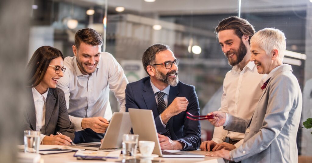 A group of diverse financial professionals smiling and collaborating around a laptop in a modern office, representing a strong sense of community and teamwork.