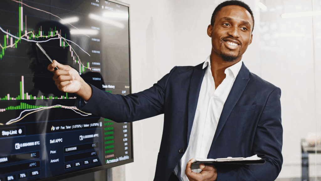 A smiling financial advisor in a suit points to a digital screen displaying market charts and trading data while holding a clipboard, illustrating investment analysis and client engagement.